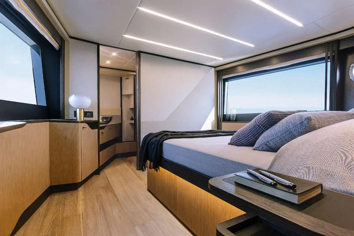 Modern yacht bedroom with large windows, wooden furnishings, a bedding setup, and a bedside table with a notebook and pens.